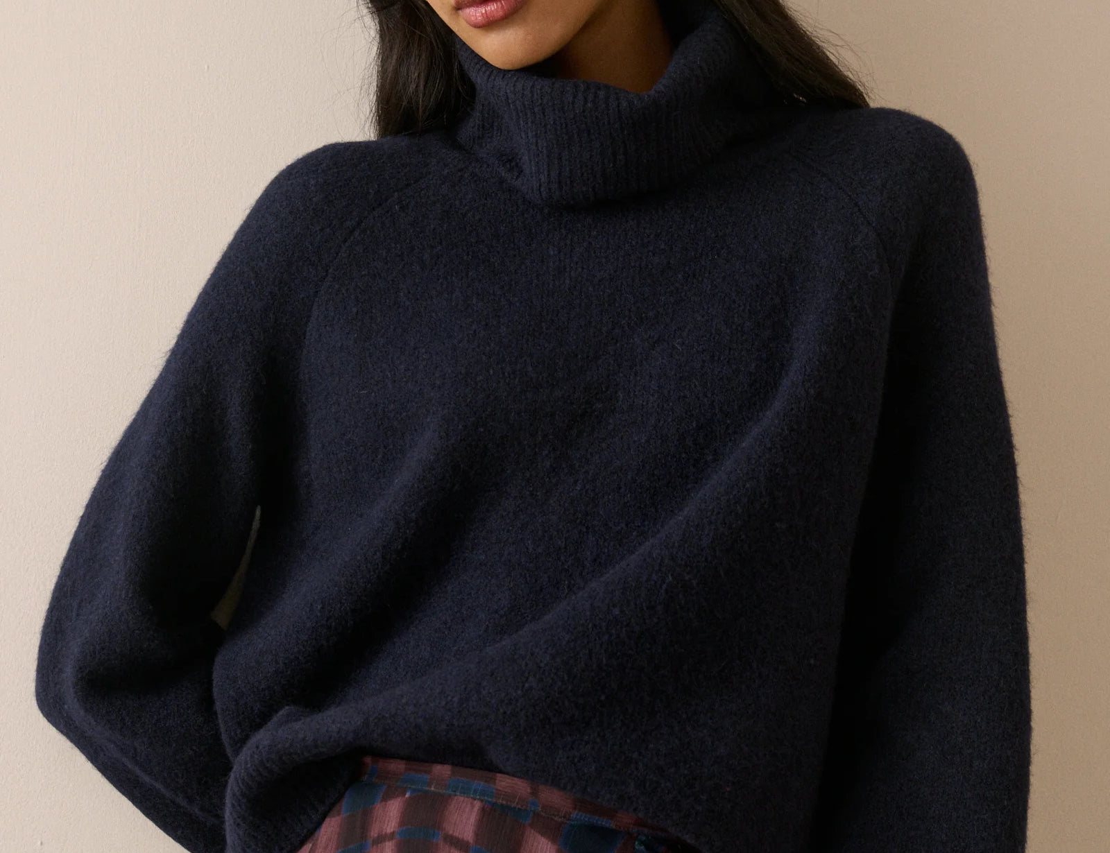 A woman wearing a dark navy blue turtleneck sweater and a plaid skirt, photographed against a plain beige background.
