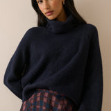 A woman wearing a dark navy blue turtleneck sweater and a plaid skirt, photographed against a plain beige background.