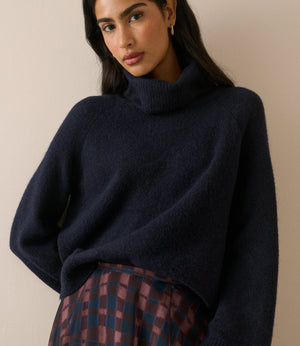 A woman wearing a dark navy blue turtleneck sweater and a plaid skirt, photographed against a plain beige background.