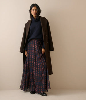 A woman stands against a plain beige studio background wearing a long dark brown coat over a navy turtleneck and a long navy skirt with a red and navy check pattern.
