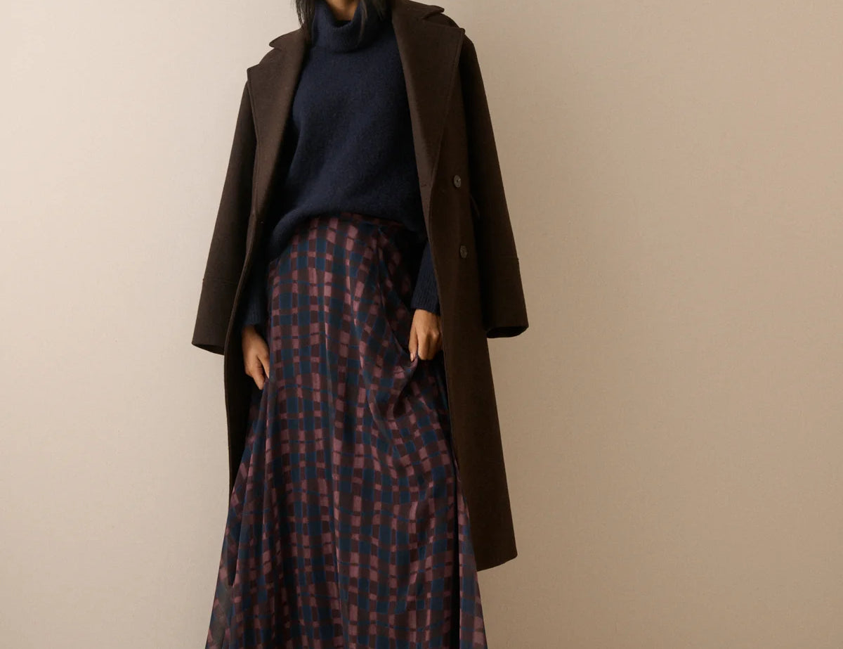 A woman stands against a plain beige studio background wearing a long dark brown coat over a navy turtleneck and a long navy skirt with a red and navy check pattern.