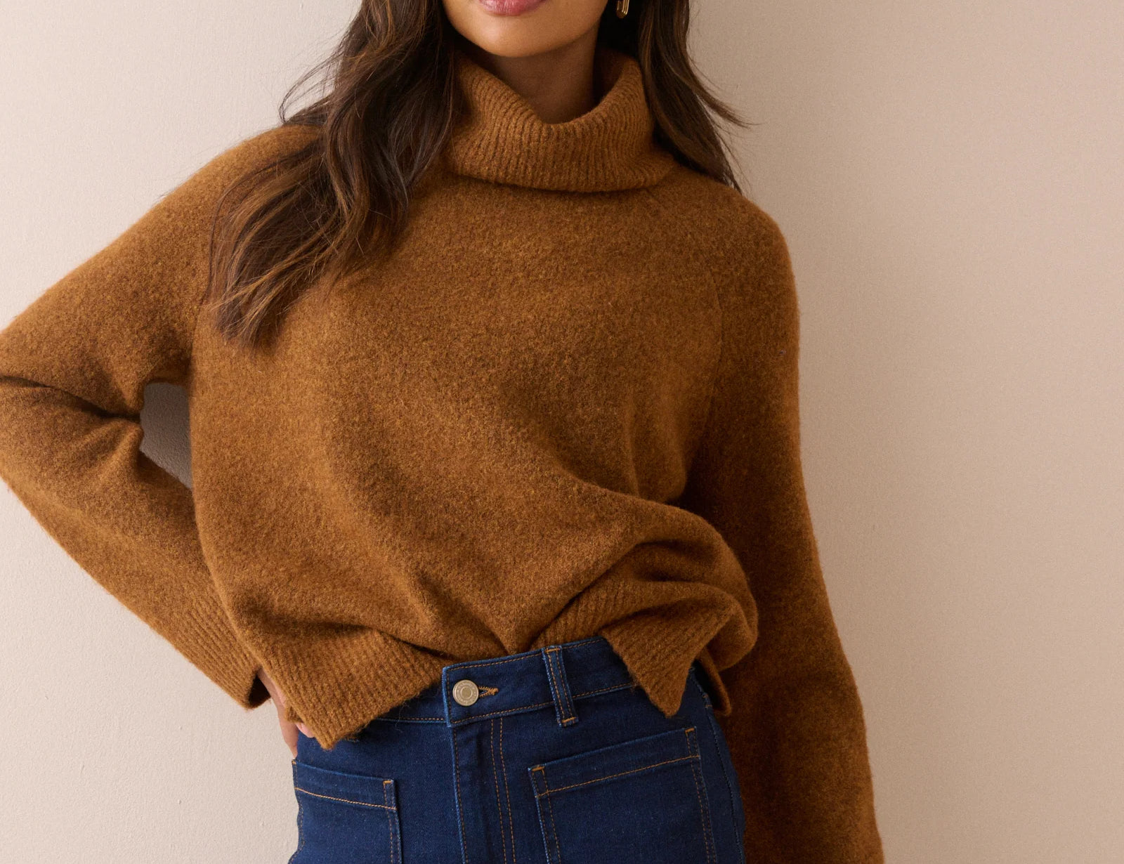 A woman wearing a warm camel brown knit turtleneck sweater and dark blue jeans, posing against a solid beige wall.