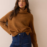A woman wearing a warm camel brown knit turtleneck sweater and dark blue jeans, posing against a solid beige wall.