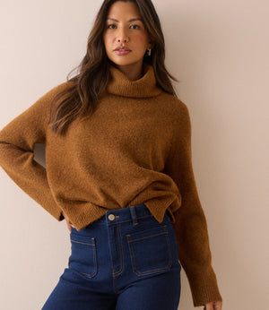 A woman wearing a warm camel brown knit turtleneck sweater and dark blue jeans, posing against a solid beige wall.