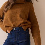 A woman wearing a fuzzy, oversized brown turtleneck sweater and dark blue jeans, shown from the chest down against a plain beige background.