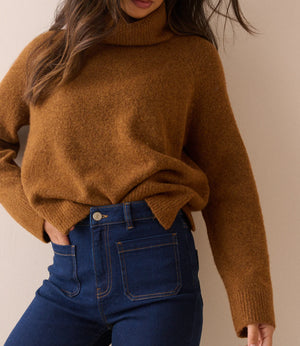 A woman wearing a fuzzy, oversized brown turtleneck sweater and dark blue jeans, shown from the chest down against a plain beige background.