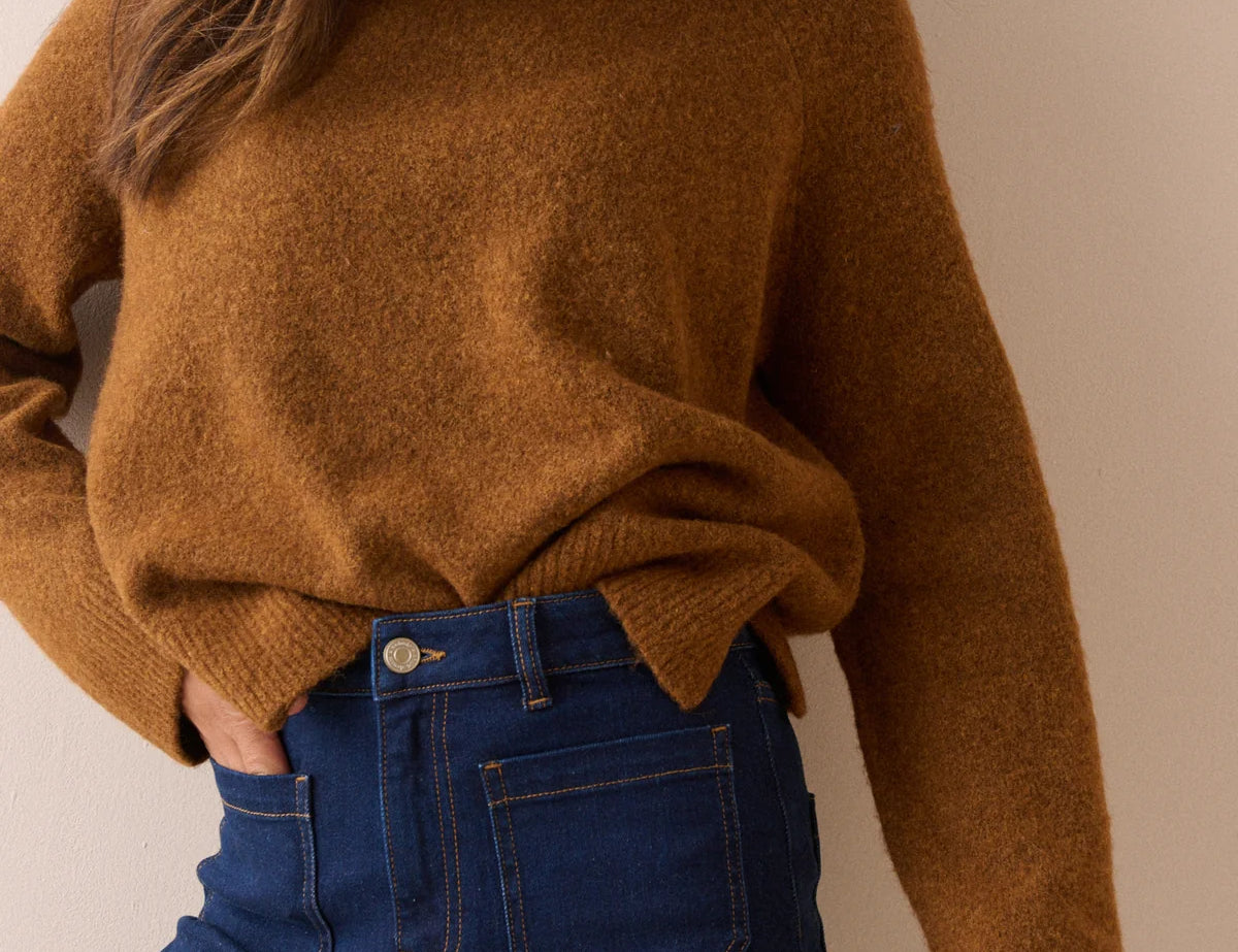 A woman wearing a fuzzy, oversized brown turtleneck sweater and dark blue jeans, shown from the chest down against a plain beige background.