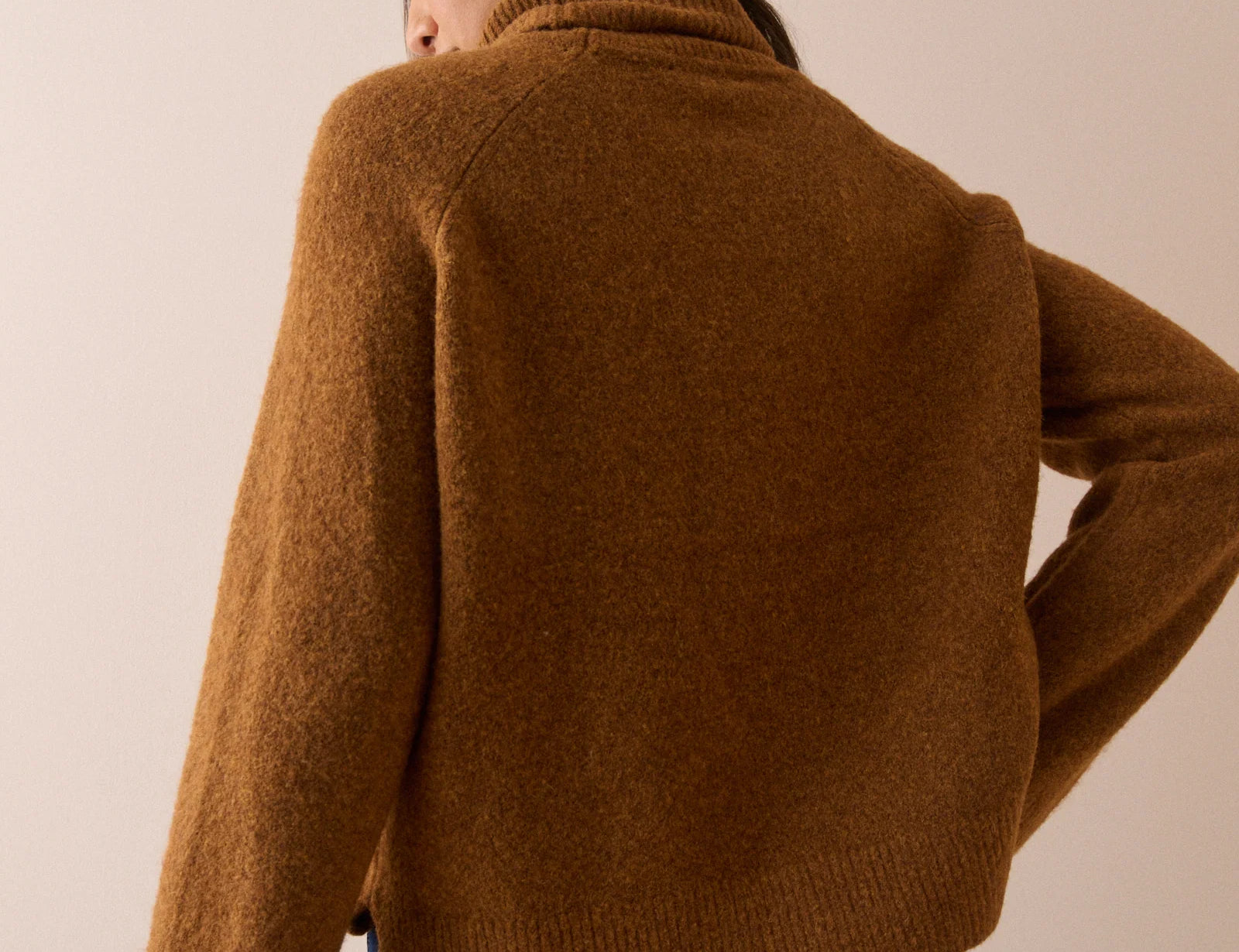 A woman shown from the back wearing a cropped, long-sleeve knit sweater in a warm camel brown color, paired with dark blue jeans, against a solid beige studio background.