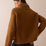A woman shown from the back wearing a cropped, long-sleeve knit sweater in a warm camel brown color, paired with dark blue jeans, against a solid beige studio background.