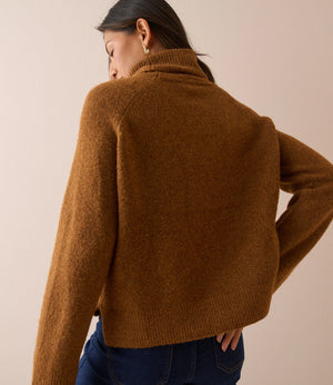 A woman shown from the back wearing a cropped, long-sleeve knit sweater in a warm camel brown color, paired with dark blue jeans, against a solid beige studio background.