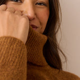 Close-up of a woman wearing a warm brown ribbed turtleneck sweater, with her hand near her face against a plain beige background.