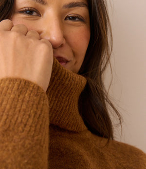Close-up of a woman wearing a warm brown ribbed turtleneck sweater, with her hand near her face against a plain beige background.