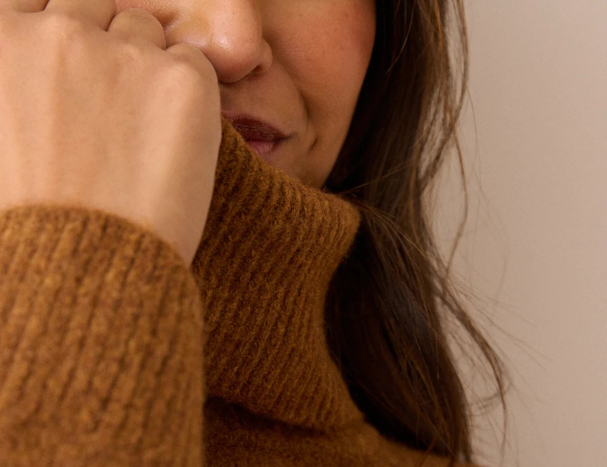 Close-up of a woman wearing a warm brown ribbed turtleneck sweater, with her hand near her face against a plain beige background.