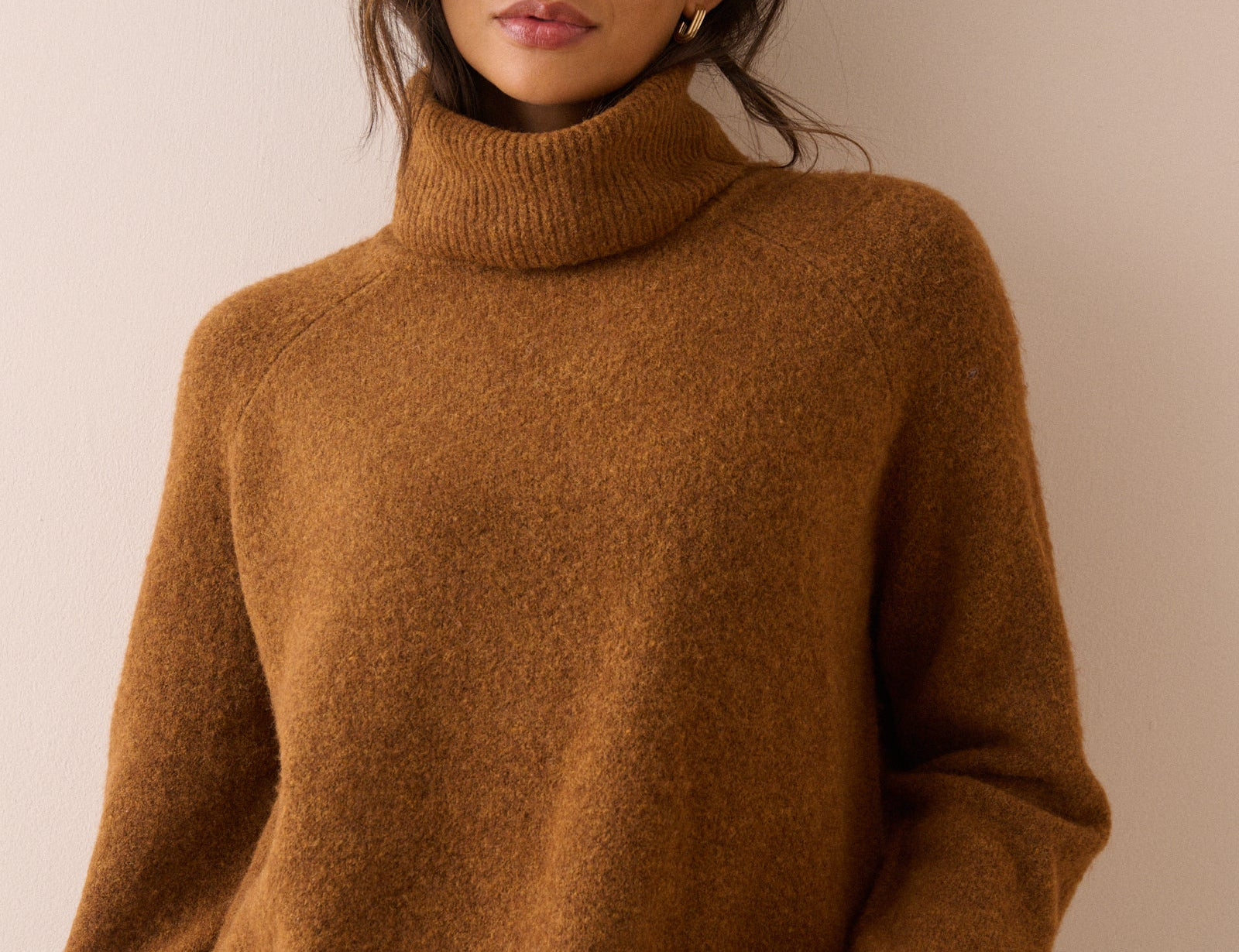 A woman wearing a cozy, textured brown turtleneck sweater and blue jeans, standing against a plain beige wall.