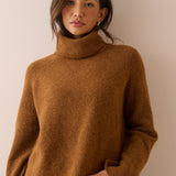A woman wearing a cozy, textured brown turtleneck sweater and blue jeans, standing against a plain beige wall.