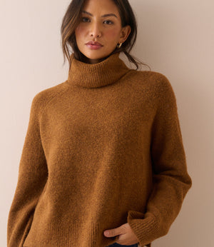 A woman wearing a cozy, textured brown turtleneck sweater and blue jeans, standing against a plain beige wall.
