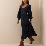 A woman modeling a navy blue midi dress with long puff sleeves, a square neckline, and a tiered skirt, paired with brown knee-high heeled boots, in a minimalist beige studio setting.