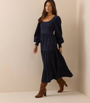 A woman modeling a navy blue midi dress with long puff sleeves, a square neckline, and a tiered skirt, paired with brown knee-high heeled boots, in a minimalist beige studio setting.
