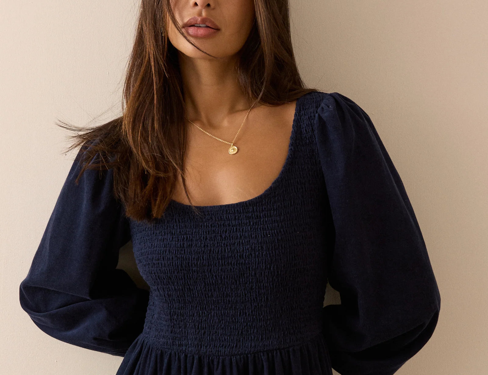 A woman wearing a dark navy blue smocked dress with long puff sleeves, standing against a plain beige wall.