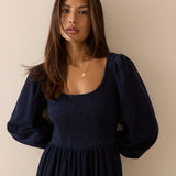 A woman wearing a dark navy blue smocked dress with long puff sleeves, standing against a plain beige wall.