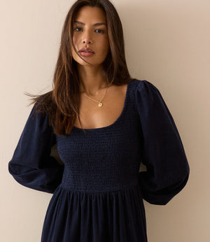 A woman wearing a dark navy blue smocked dress with long puff sleeves, standing against a plain beige wall.