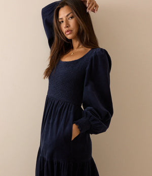 A woman wearing a dark navy blue smocked dress with long puff sleeves and a tiered skirt, posing against a solid beige background.