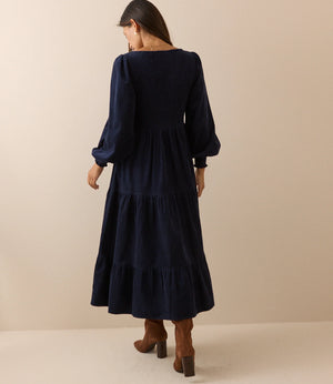 A woman wearing a dark navy blue tiered midi dress with long balloon sleeves, shown from the back against a solid beige studio background.