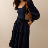A woman wearing a navy blue tiered maxi dress with voluminous puff sleeves and side pockets, carrying a brown leather shoulder bag, set against a plain beige studio background.