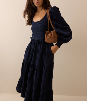 A woman wearing a navy blue tiered maxi dress with voluminous puff sleeves and side pockets, carrying a brown leather shoulder bag, set against a plain beige studio background.