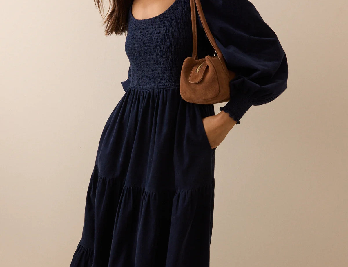 A woman wearing a navy blue tiered maxi dress with voluminous puff sleeves and side pockets, carrying a brown leather shoulder bag, set against a plain beige studio background.