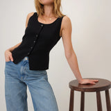Woman wearing a black sleeveless top and blue jeans standing next to a wooden stool on a plain background