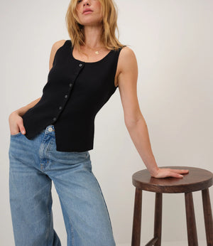 Woman wearing a black sleeveless top and blue jeans standing next to a wooden stool on a plain background
