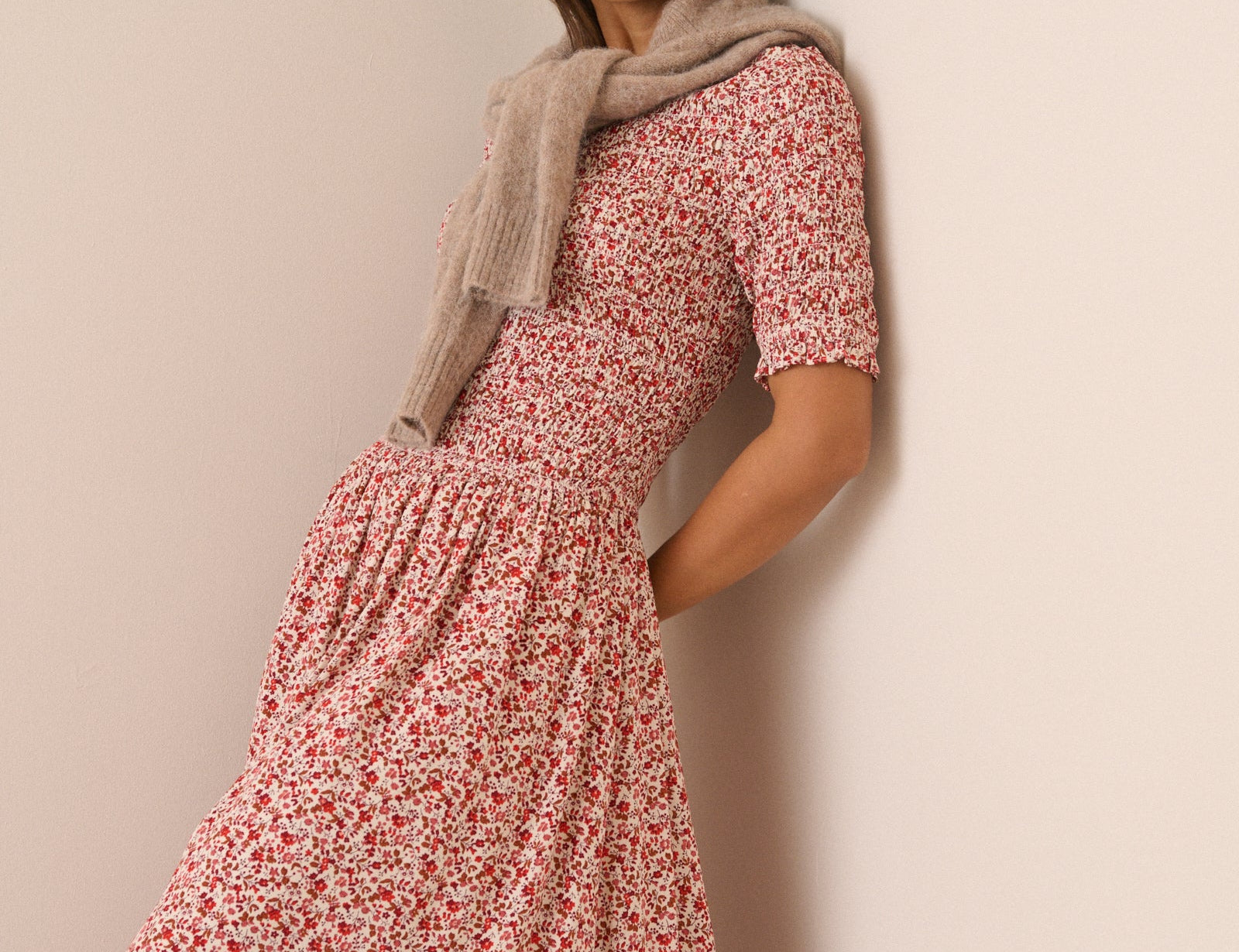 A woman wearing a short-sleeved midi dress with a red and white floral print, styled with a beige knit scarf, leaning against a plain beige wall.