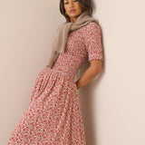 A woman wearing a short-sleeved midi dress with a red and white floral print, styled with a beige knit scarf, leaning against a plain beige wall.