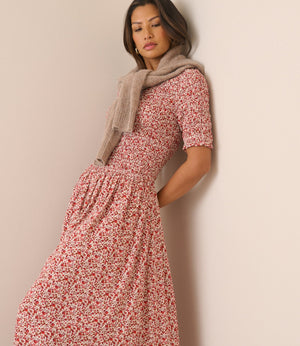 A woman wearing a short-sleeved midi dress with a red and white floral print, styled with a beige knit scarf, leaning against a plain beige wall.