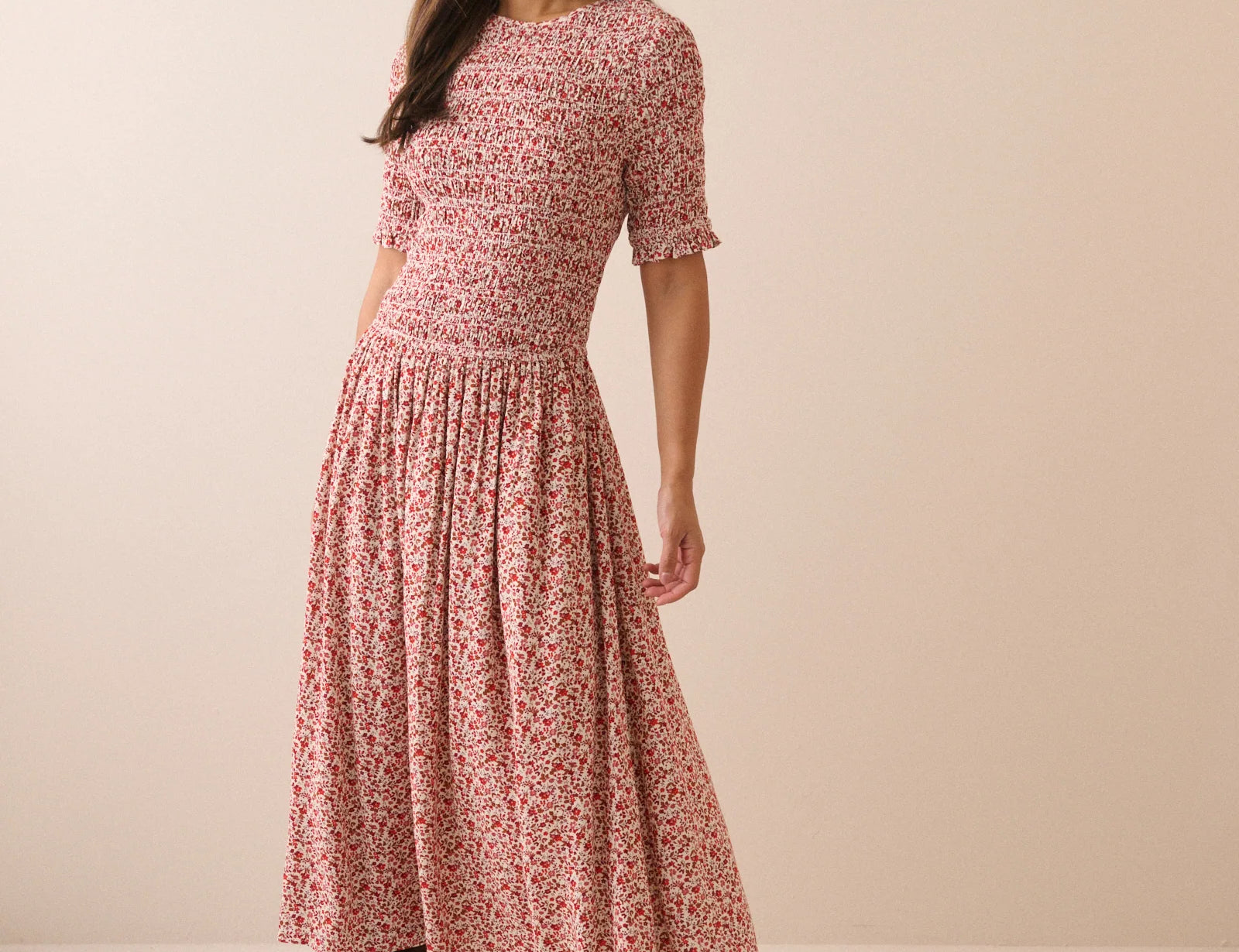 A woman modeling a short-sleeve midi dress with a dense red and pink floral print on a light beige base, paired with dark brown ankle boots, against a solid beige studio background.