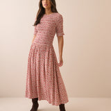 A woman modeling a short-sleeve midi dress with a dense red and pink floral print on a light beige base, paired with dark brown ankle boots, against a solid beige studio background.