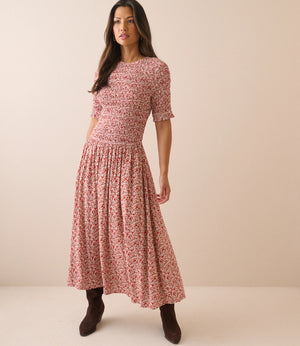 A woman modeling a short-sleeve midi dress with a dense red and pink floral print on a light beige base, paired with dark brown ankle boots, against a solid beige studio background.
