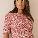 A woman wearing a short-sleeve smocked dress with a red and white floral print, photographed against a plain beige wall.