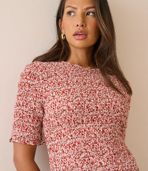 A woman wearing a short-sleeve smocked dress with a red and white floral print, photographed against a plain beige wall.