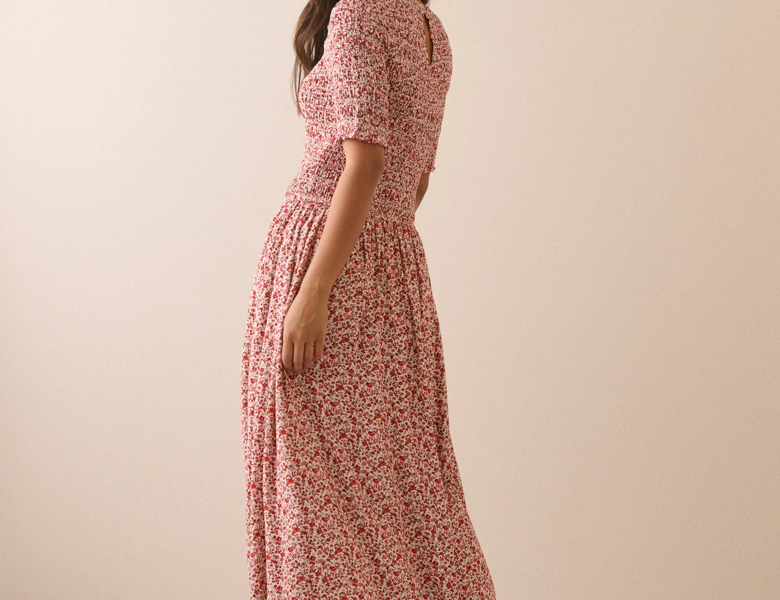 A woman modeling a short-sleeve midi dress with a pink and red floral pattern, shown from a side-back angle against a plain beige studio backdrop.