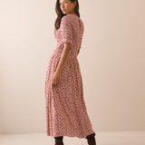 A woman modeling a short-sleeve midi dress with a pink and red floral pattern, shown from a side-back angle against a plain beige studio backdrop.
