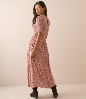 A woman modeling a short-sleeve midi dress with a pink and red floral pattern, shown from a side-back angle against a plain beige studio backdrop.