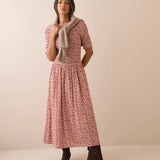 A woman modeling a short-sleeve midi dress with a red and pink floral print, styled with a beige knit cardigan and dark brown ankle boots against a solid beige studio background.