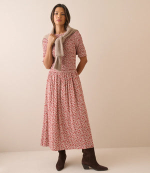 A woman modeling a short-sleeve midi dress with a red and pink floral print, styled with a beige knit cardigan and dark brown ankle boots against a solid beige studio background.