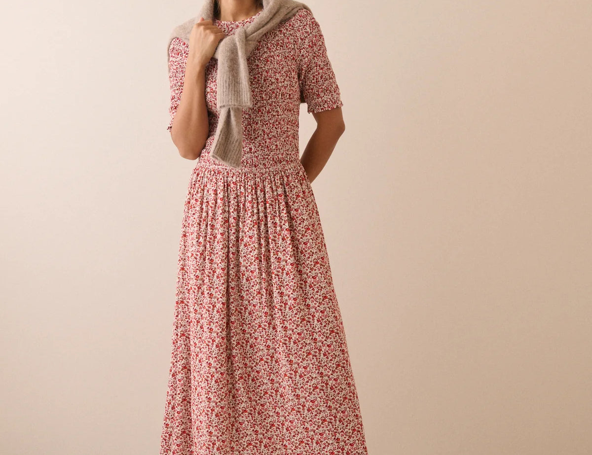 A woman modeling a short-sleeve midi dress with a red and pink floral print, styled with a beige knit cardigan and dark brown ankle boots against a solid beige studio background.