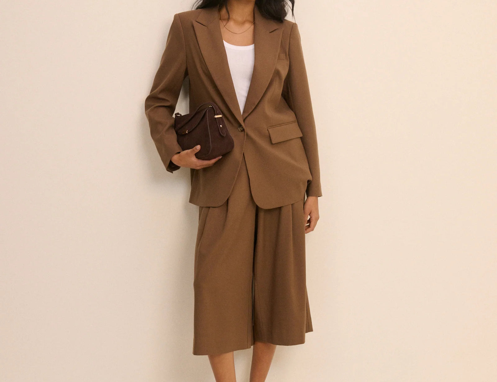 A woman models a brown tailored suit set consisting of a blazer and matching cropped trousers, styled with a white top, a dark brown handbag, and dark brown heels against a plain beige studio background.