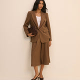 A woman models a brown tailored suit set consisting of a blazer and matching cropped trousers, styled with a white top, a dark brown handbag, and dark brown heels against a plain beige studio background.