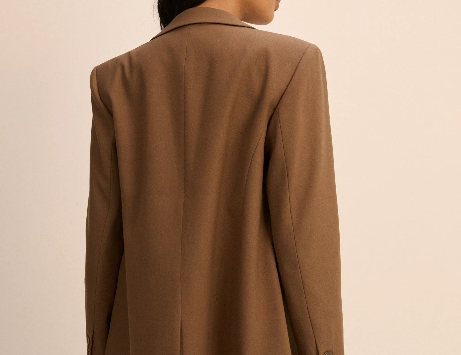 A woman shown from the back wearing an oversized brown blazer with blue jeans, photographed against a plain beige studio background.