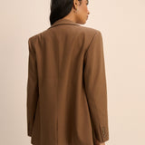 A woman shown from the back wearing an oversized brown blazer with blue jeans, photographed against a plain beige studio background.